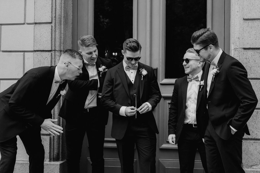 Five groomsmen in black suits examine something together outside in black and white photo.
