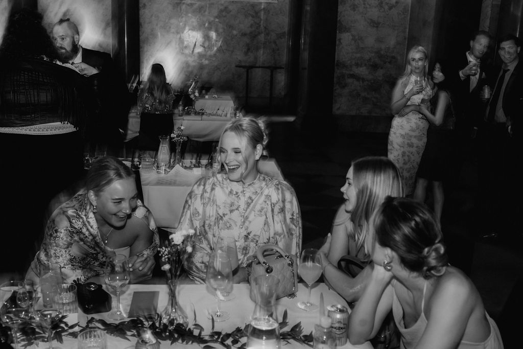Three women laugh and chat together at floral-decorated wedding reception dinner table.