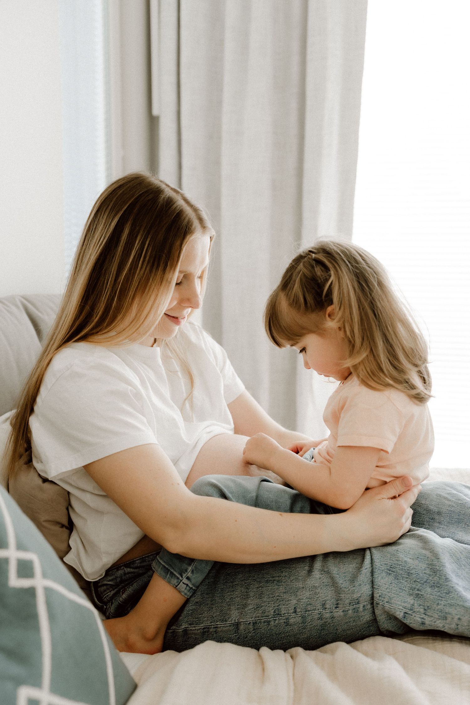 Pregnant woman in white shirt smiling at young daughter sitting on her lap on sofa.