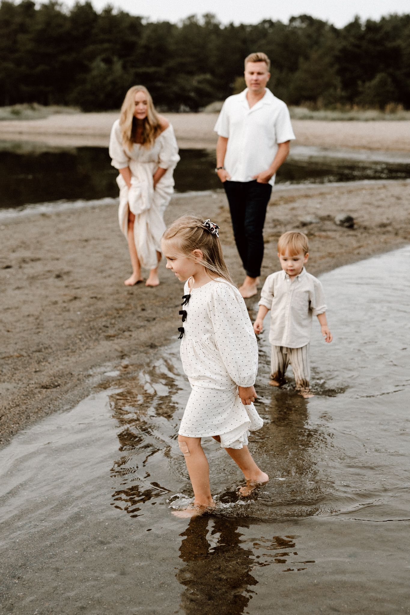 Family of four wading in shallow water on a sandy beach, parents watching two young children splash and play.