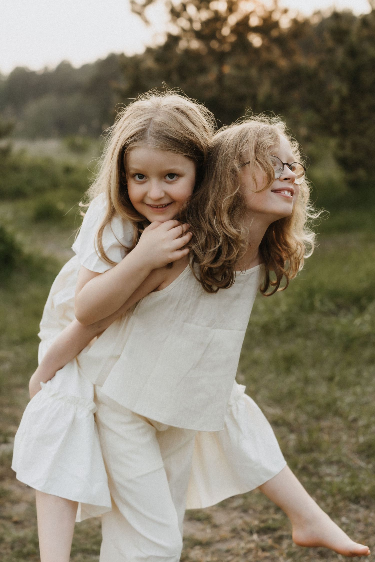 Two young sisters in white outfits smile and piggyback in a sunlit outdoor meadow.