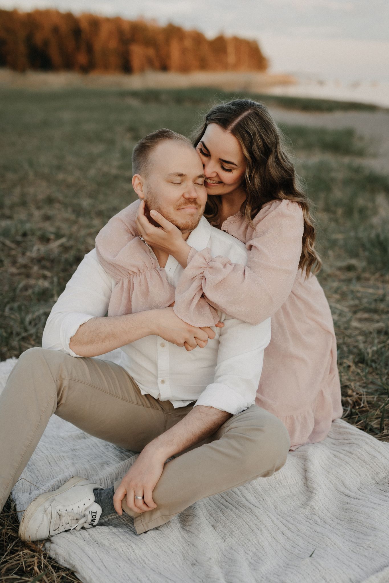 Couple sitting on blanket in field at sunset, woman in pink dress embracing man lovingly.