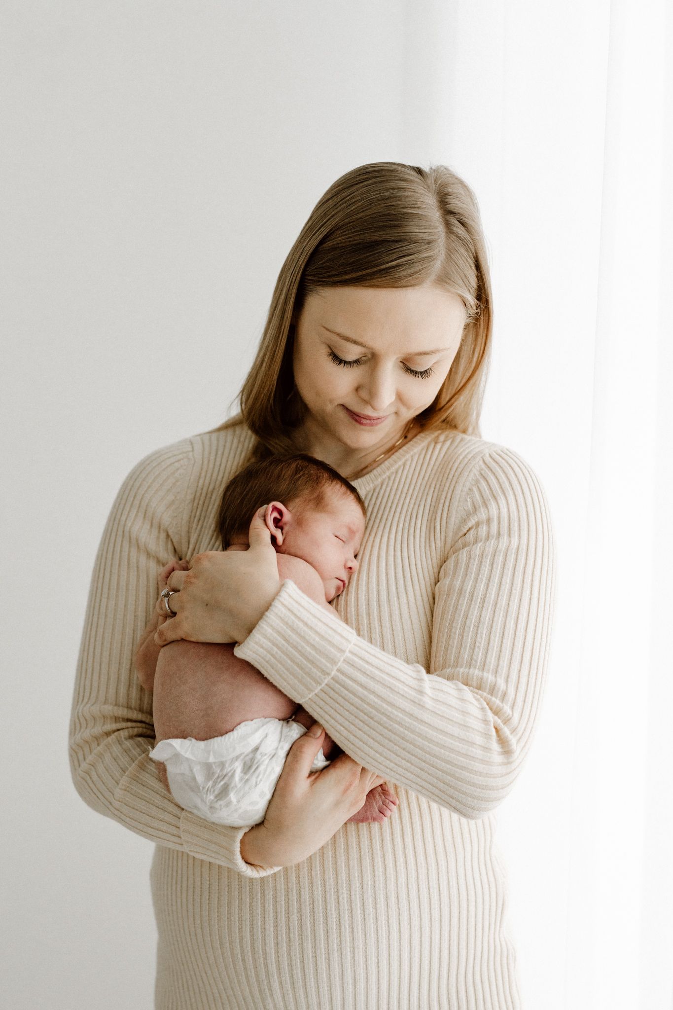Young mother in cream ribbed sweater gazes lovingly at sleeping newborn in her arms.