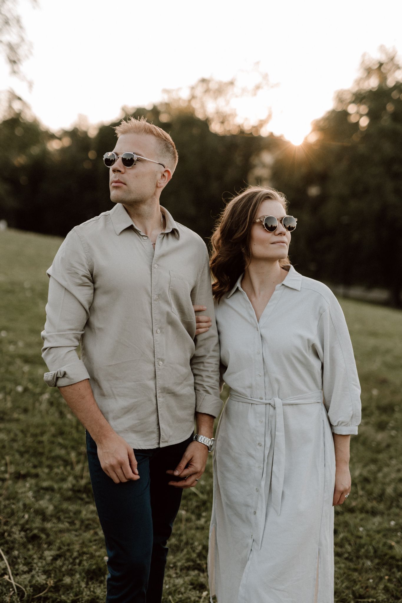 Stylish couple in matching beige outfits and sunglasses walking hand in hand at golden hour.