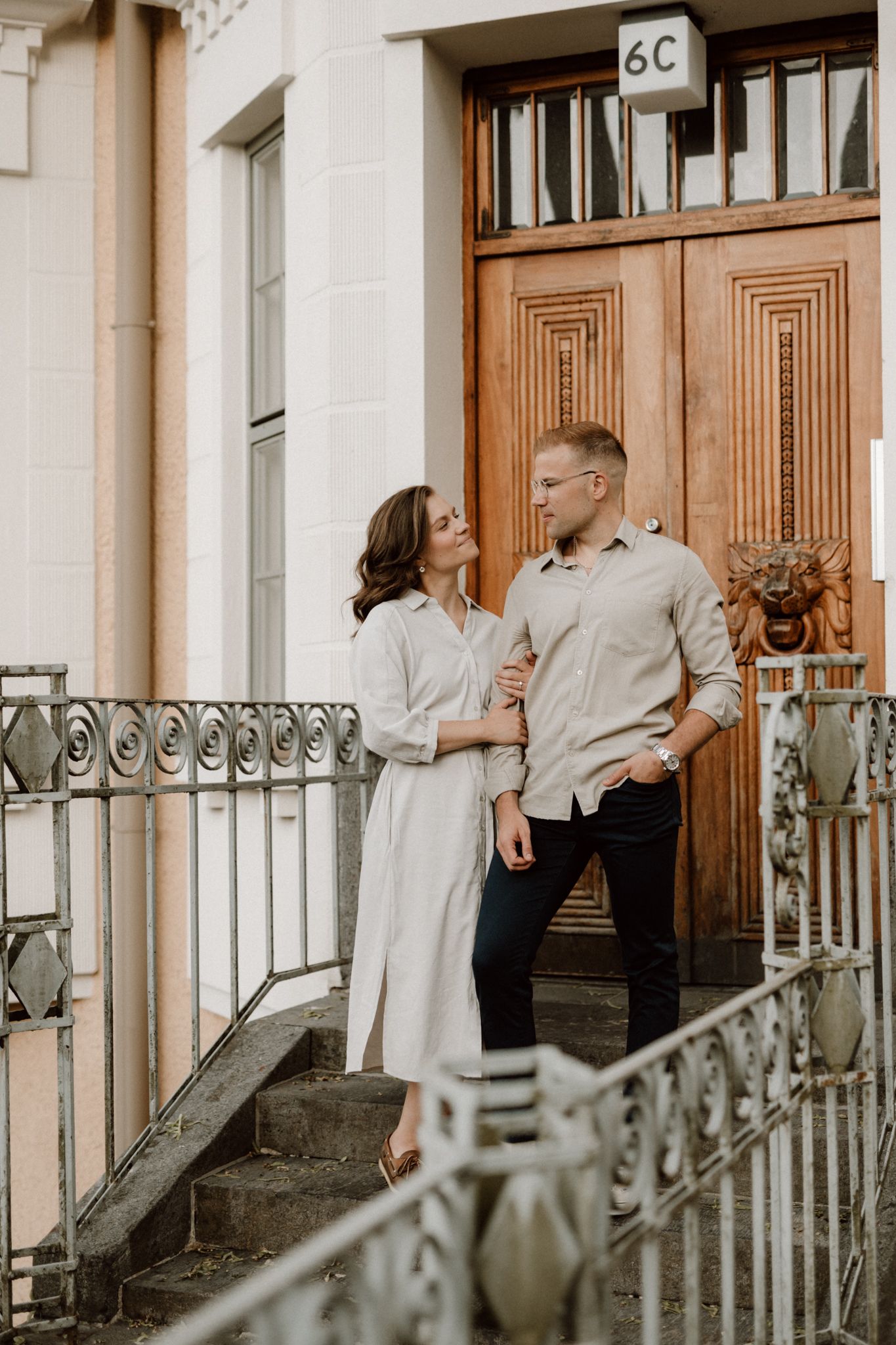 Stylish couple in neutral outfits standing together on steps before a wooden door marked 6C.