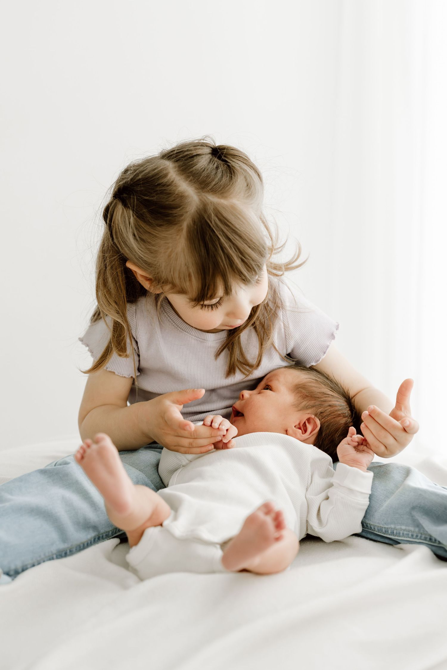 Young girl sitting on white bed looking down at crying newborn baby in her lap.