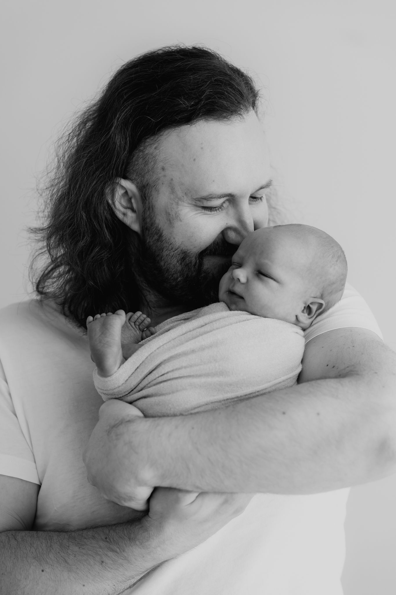 Black and white portrait of bearded father with long hair smiling at his swaddled newborn.