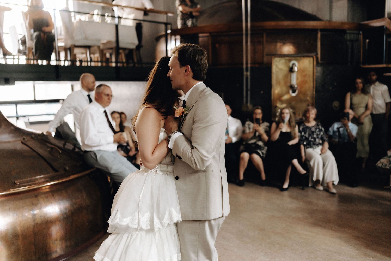 Bride and groom share their first dance beside a large copper brewery tank with guests watching.