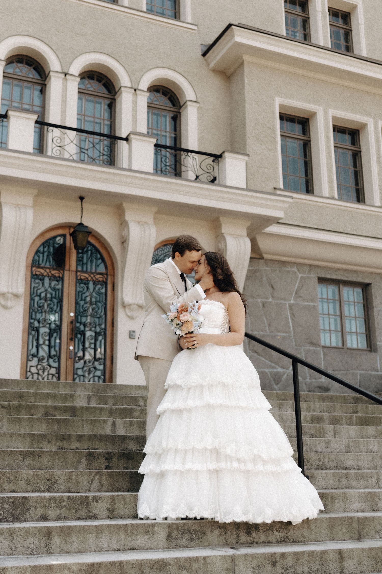 Bride and groom kissing on steps outside grand classical building, bride holding pastel bouquet.