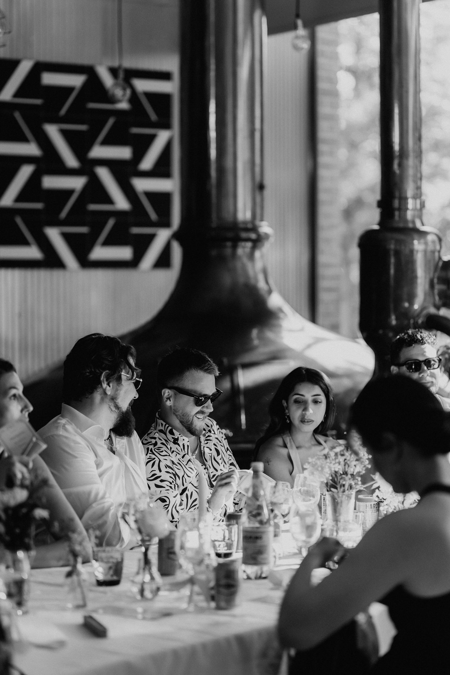 Black and white photo of wedding guests seated at a reception table near a copper still.