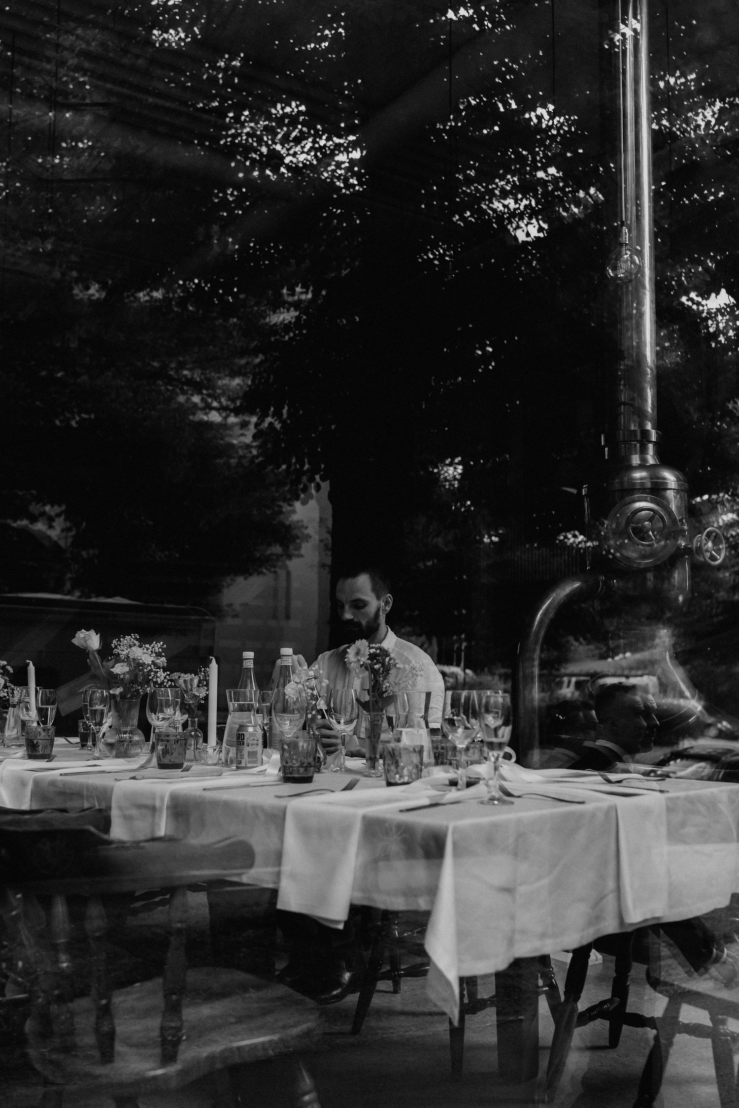 Black and white photo of a man sitting alone at an elegantly set wedding reception table near a large window.