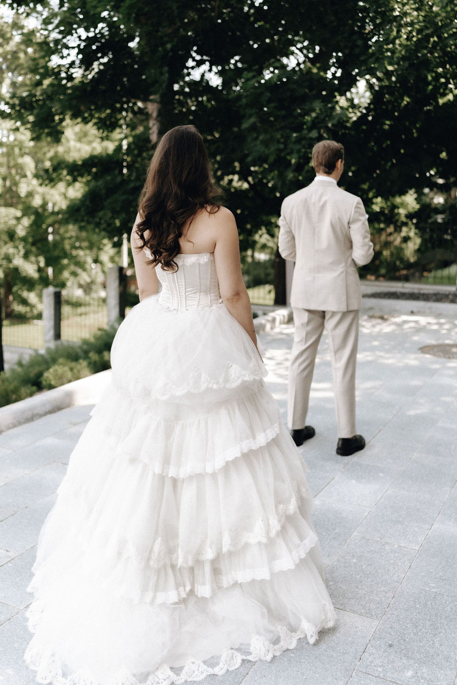 Bride in strapless layered white ballgown walking behind groom in beige suit through sunny park pathway.
