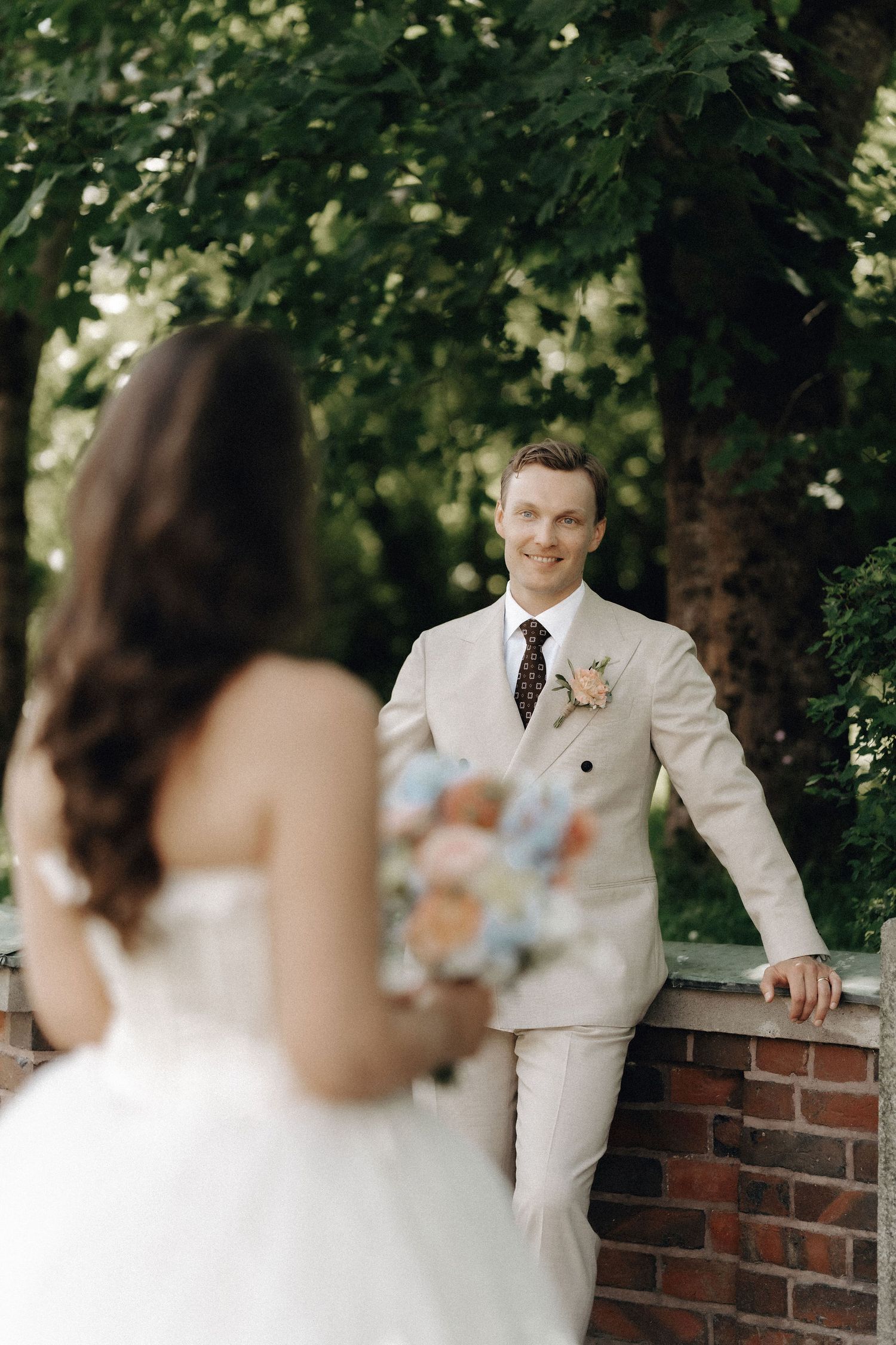 Smiling groom in beige suit leans against brick wall as bride with bouquet approaches in lush garden.