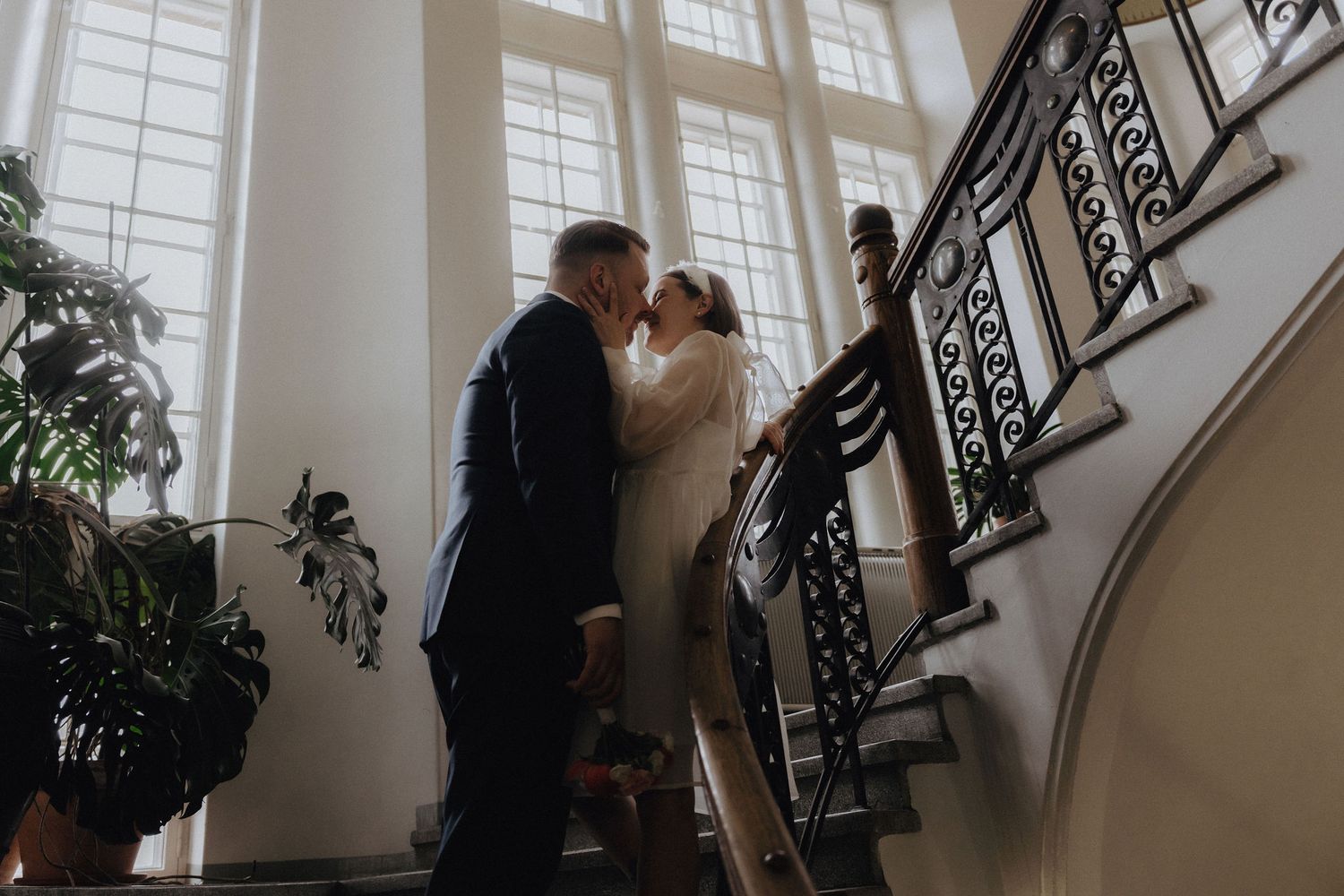 Bride and groom kiss tenderly on a grand interior staircase with ornate iron railings and tall windows.