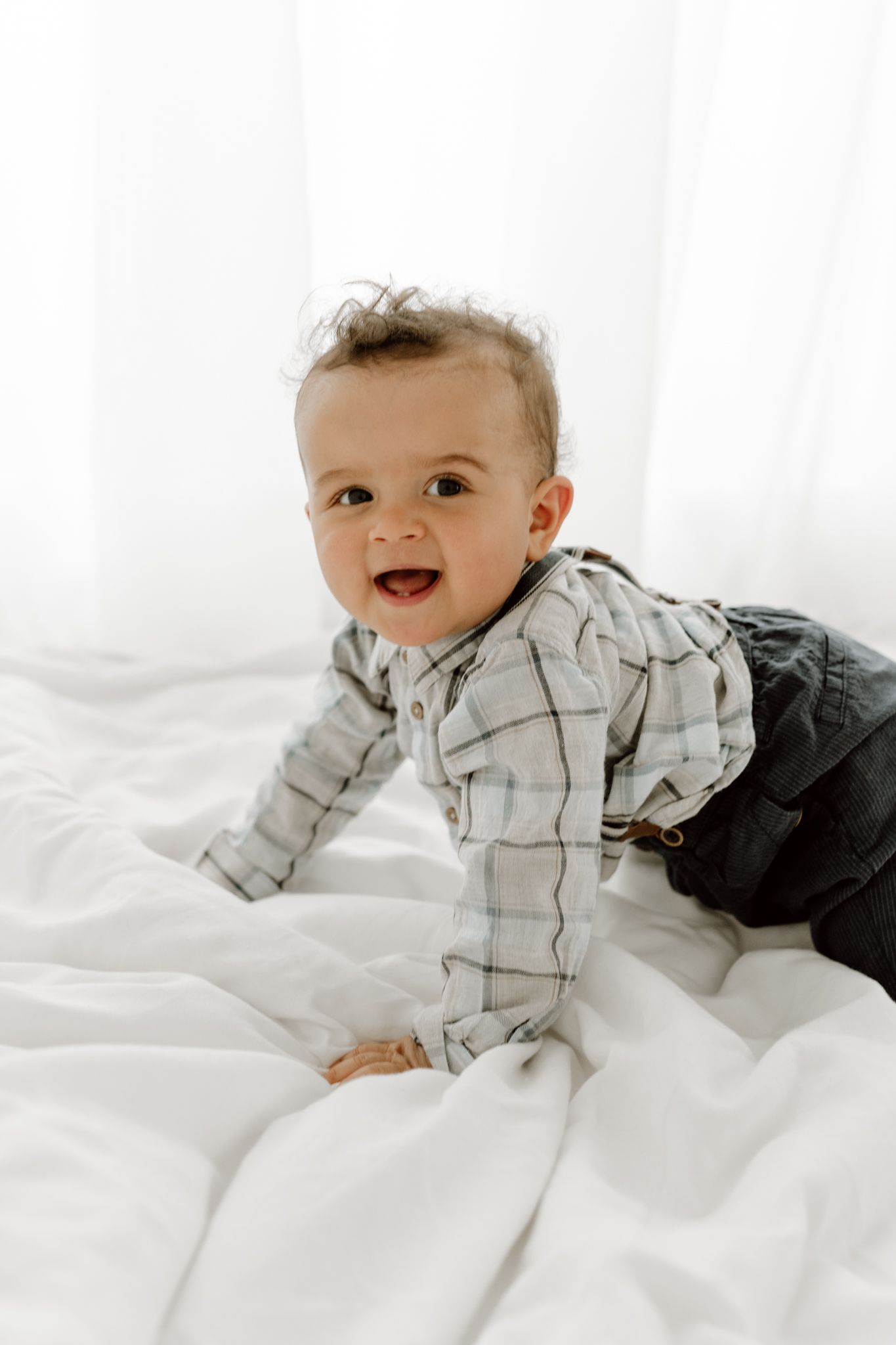 Smiling baby boy in plaid shirt and dark pants crawling on white bed linens.