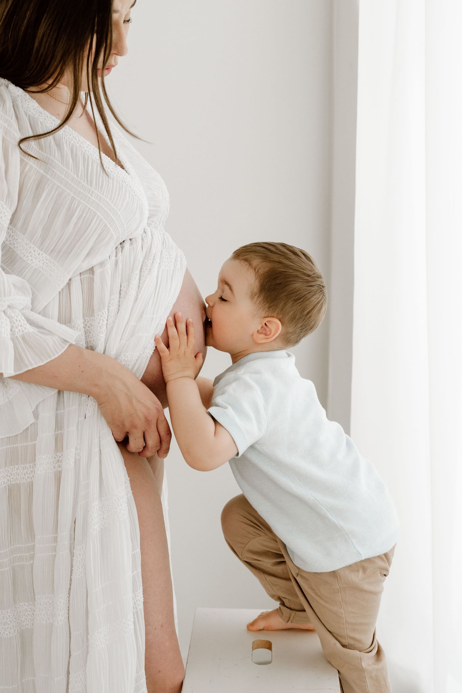 A young boy sweetly kisses his pregnant mothers belly while she stands near a bright window.