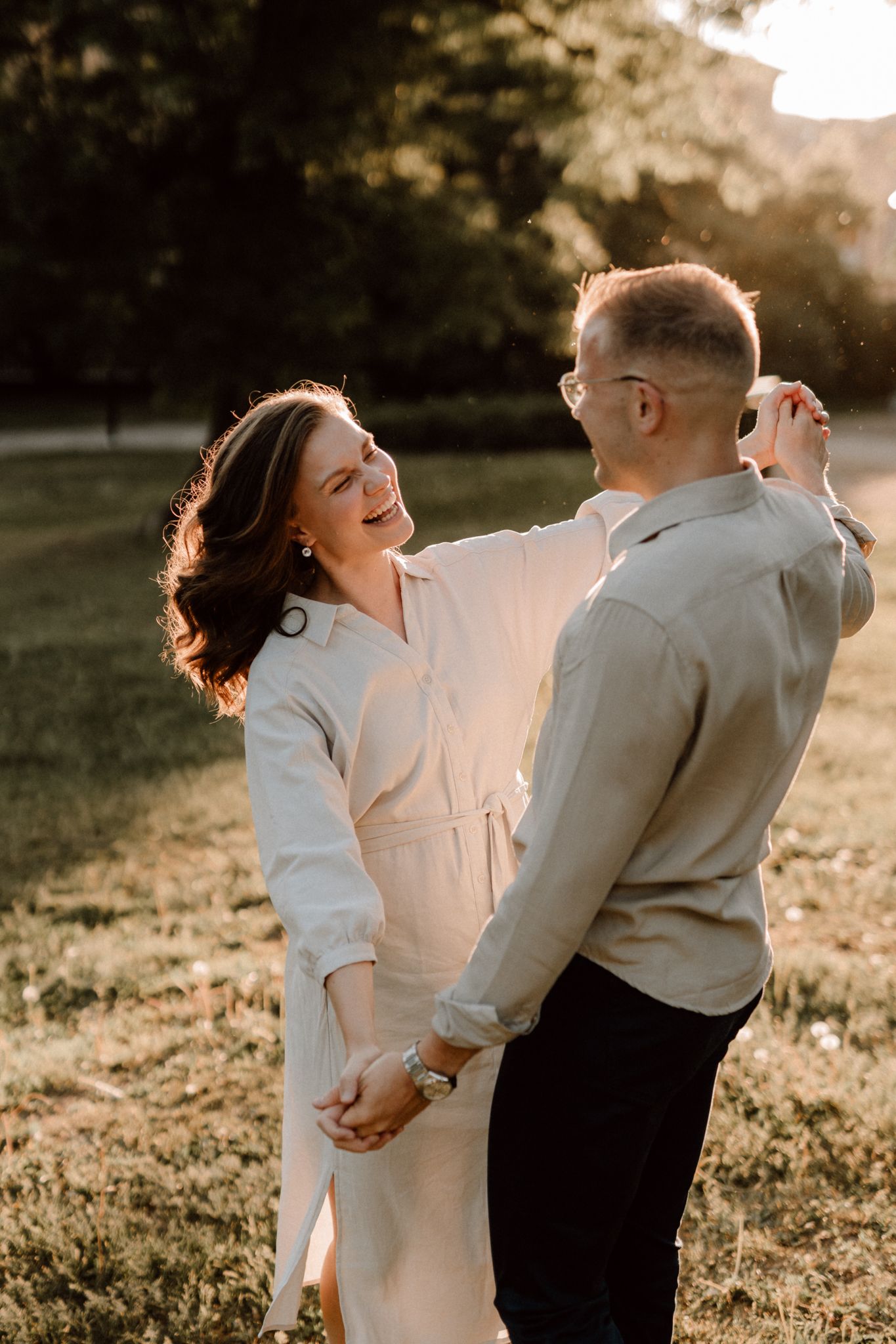 Smiling couple holding hands and dancing together in a sunlit park at golden hour.