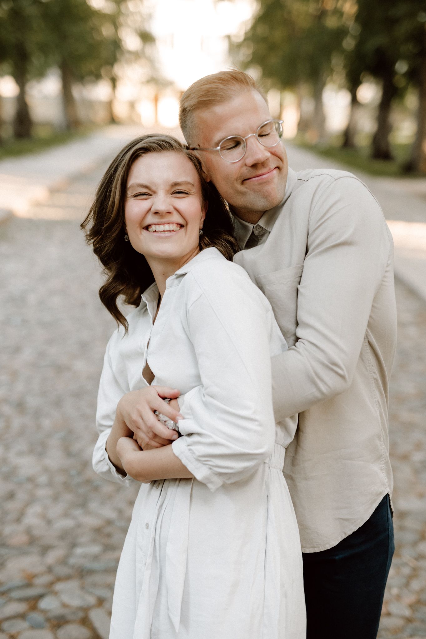 Happy couple embracing on a cobblestone path, woman laughing as man hugs her from behind at sunset.