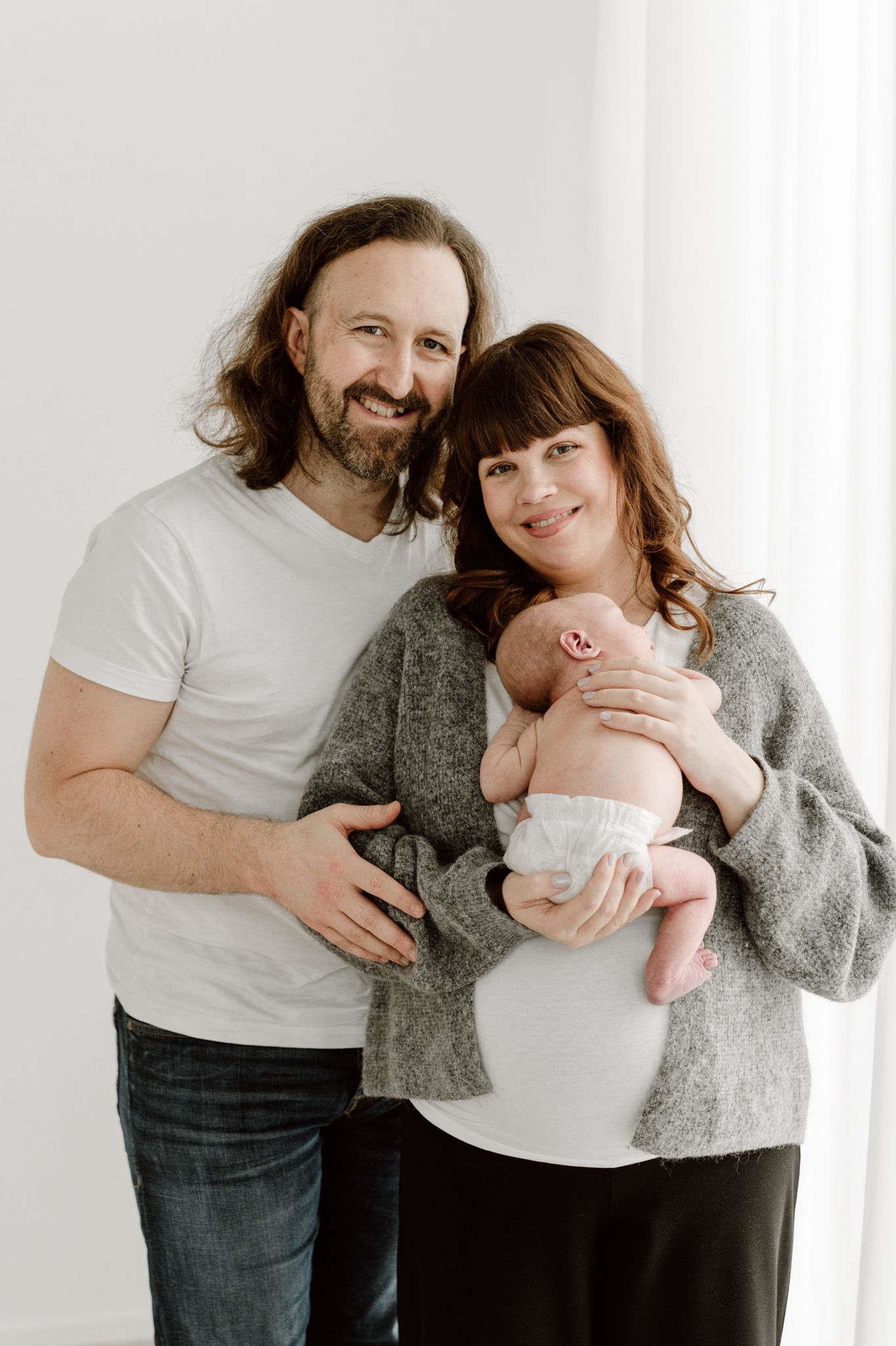 Smiling parents in a bright studio holding their newborn baby dressed in white.