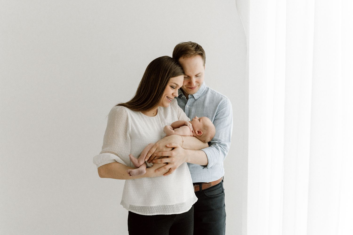 Couple in light clothing standing by window tenderly looking down at their newborn baby.