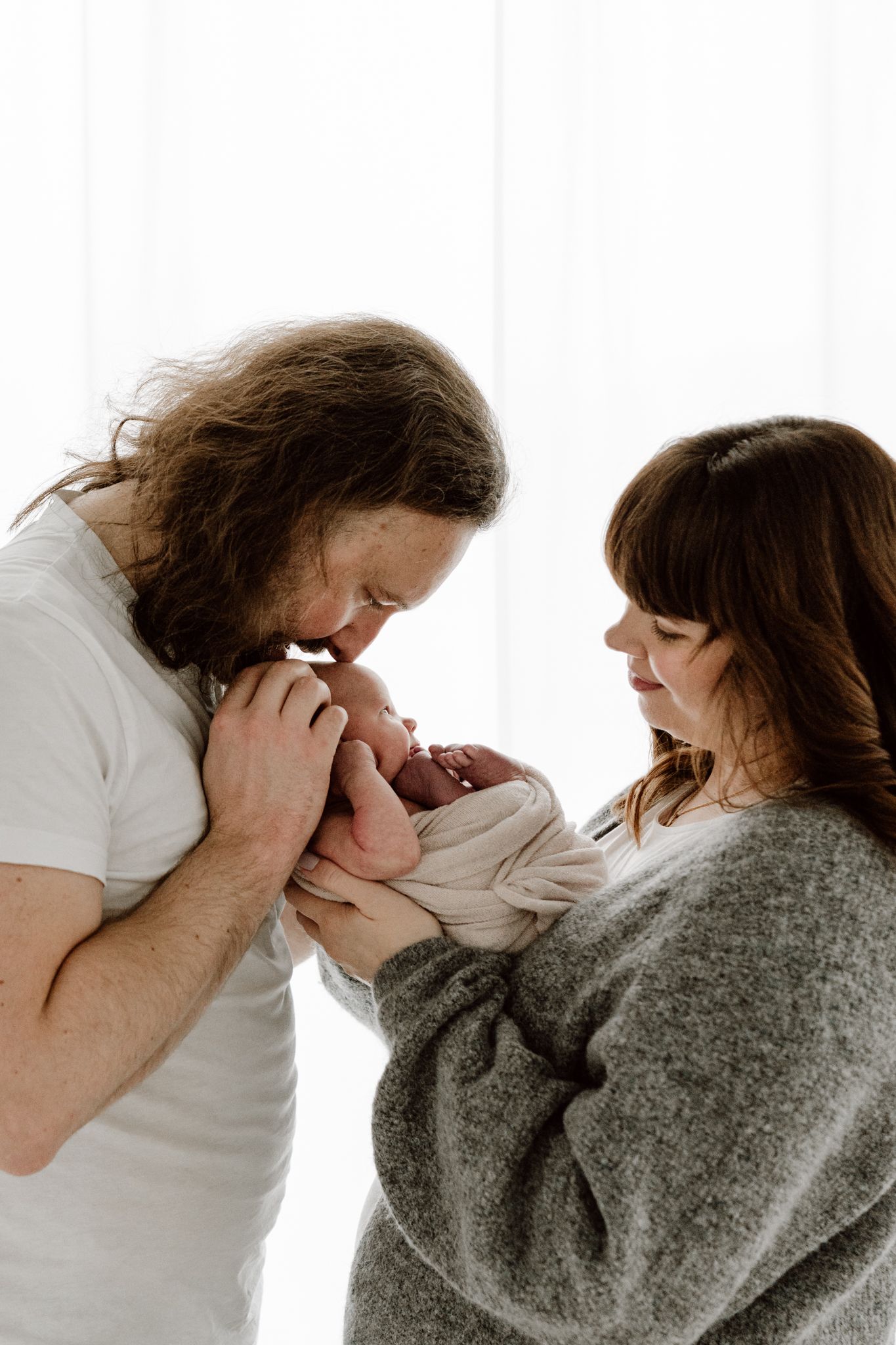 Parents leaning in together to kiss their swaddled newborn baby near a bright window.