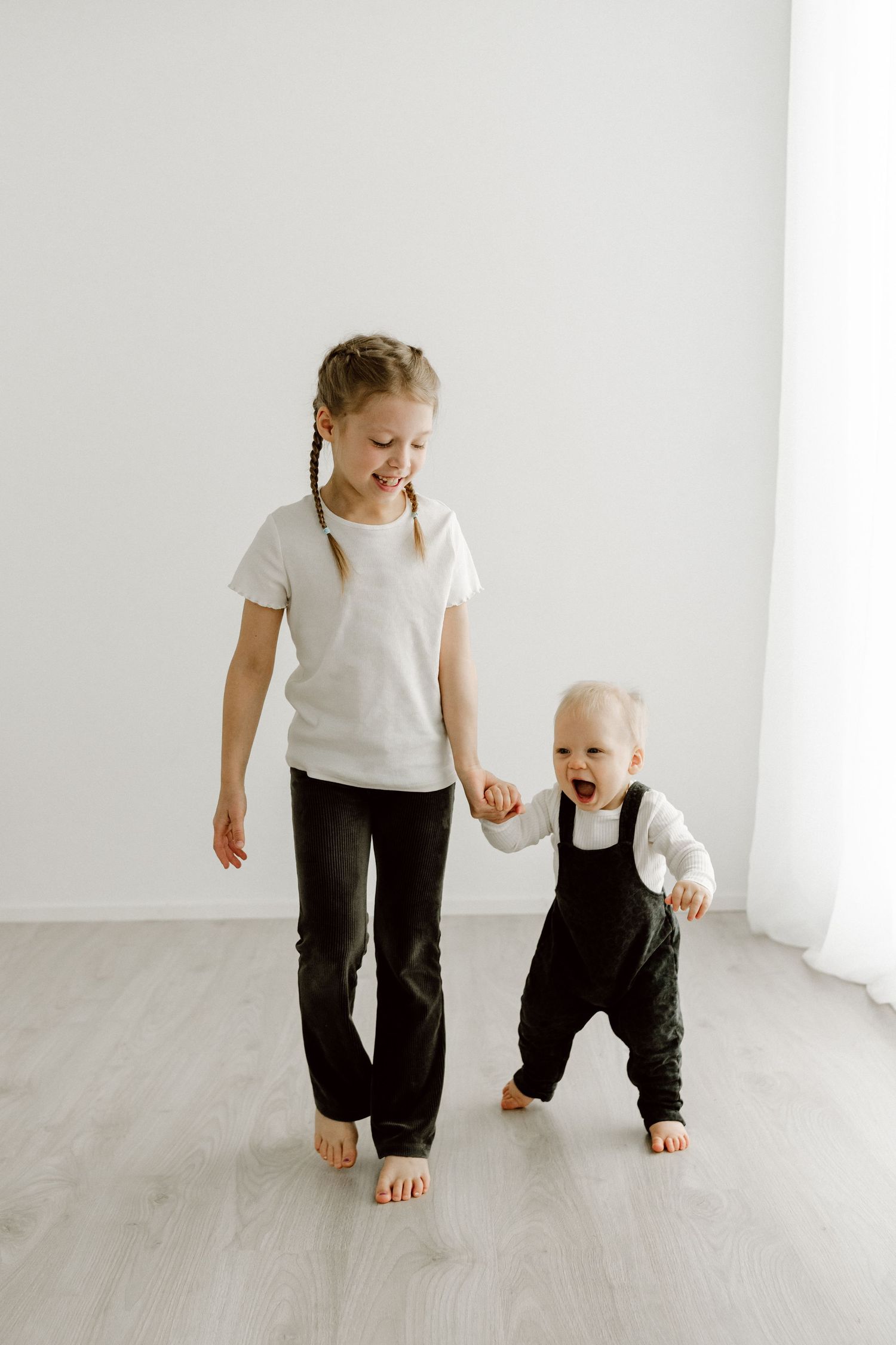 A happy older sister holds hands with her laughing toddler sibling learning to walk in a bright white room.