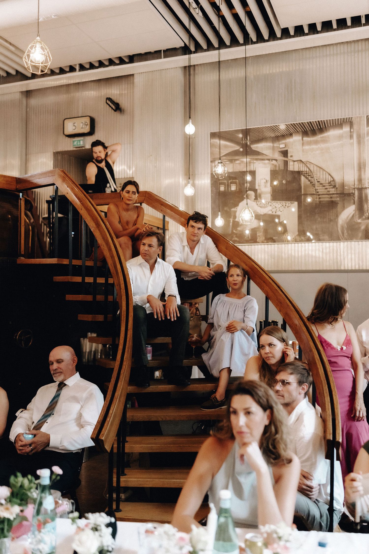 Wedding guests seated on curved wooden staircase during reception speeches.