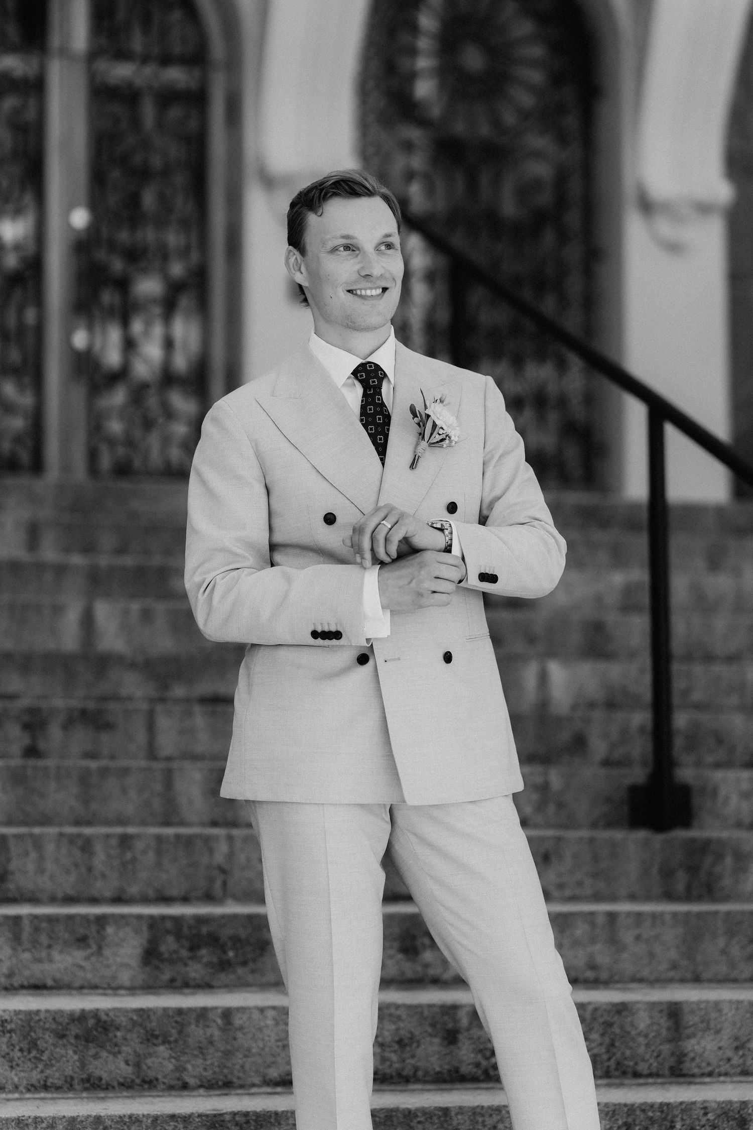 Black and white portrait of a smiling groom in a beige double-breasted suit standing on stone steps.