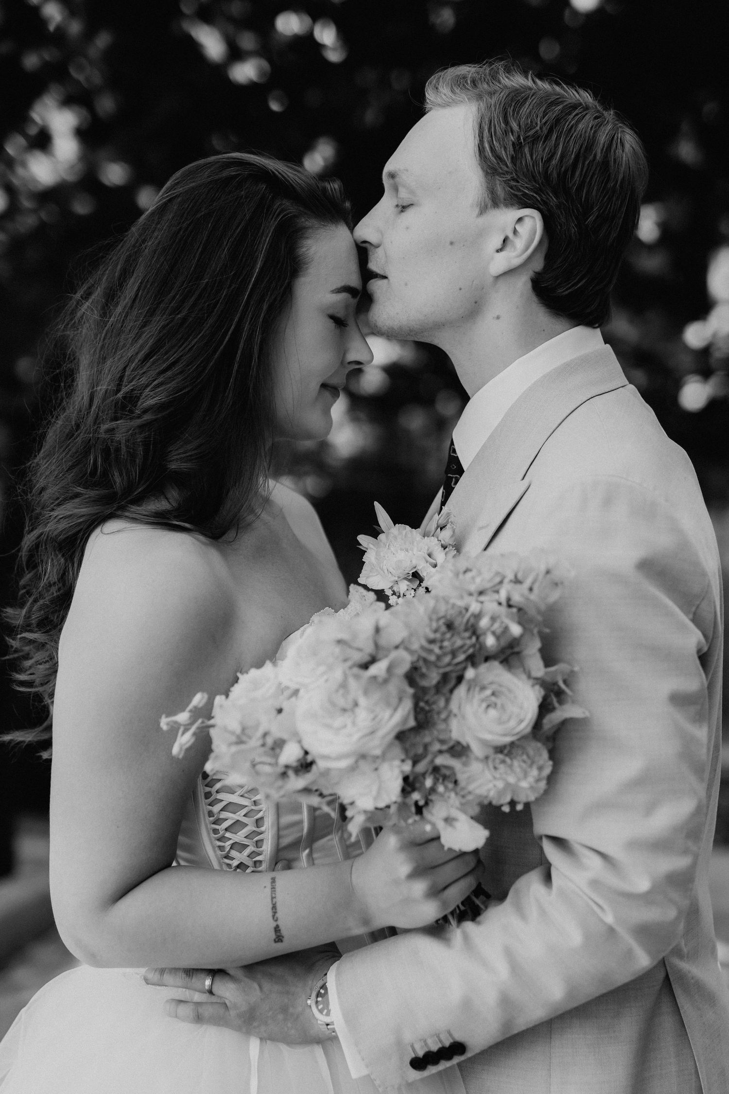 Black and white romantic photo of groom kissing bride on forehead while she holds a lush floral bouquet.