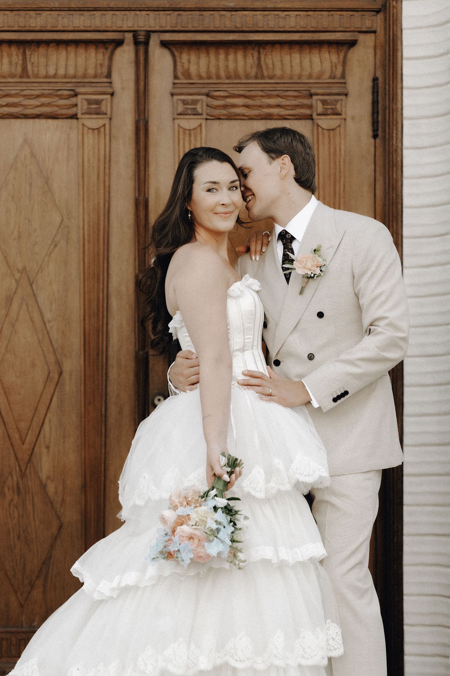 Groom whispers to smiling bride as they pose before ornate wooden doors, bride holding pastel flowers.