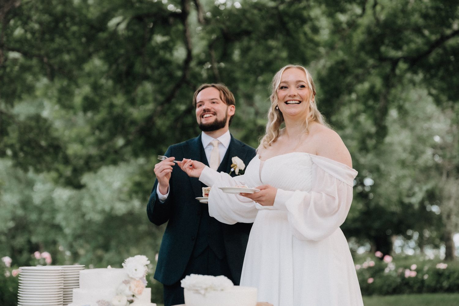 Smiling bride and groom cut elegant white wedding cake outdoors surrounded by lush green trees.