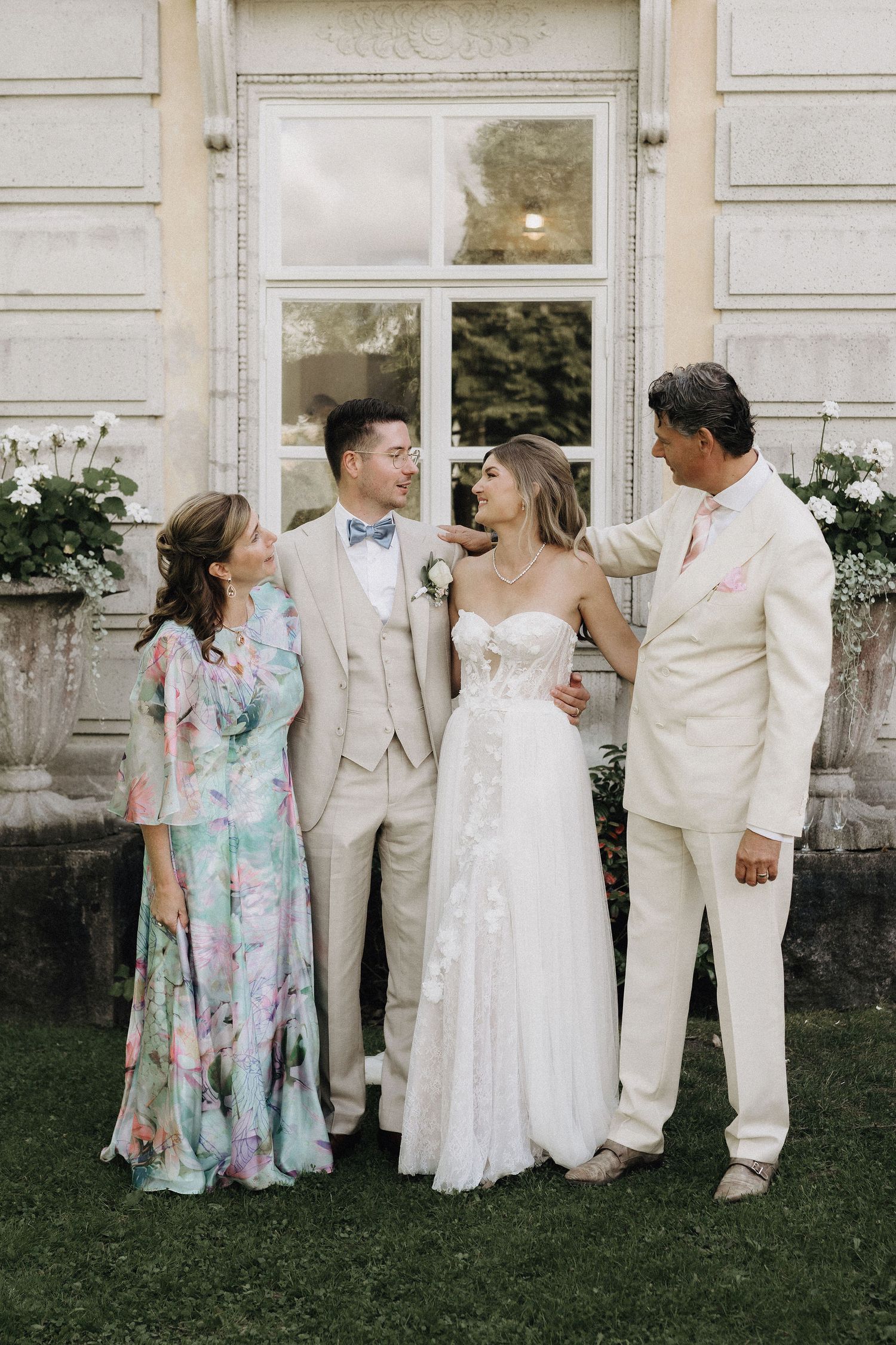 Bride in floral lace gown with groom in beige suit celebrating with parents at elegant wedding.