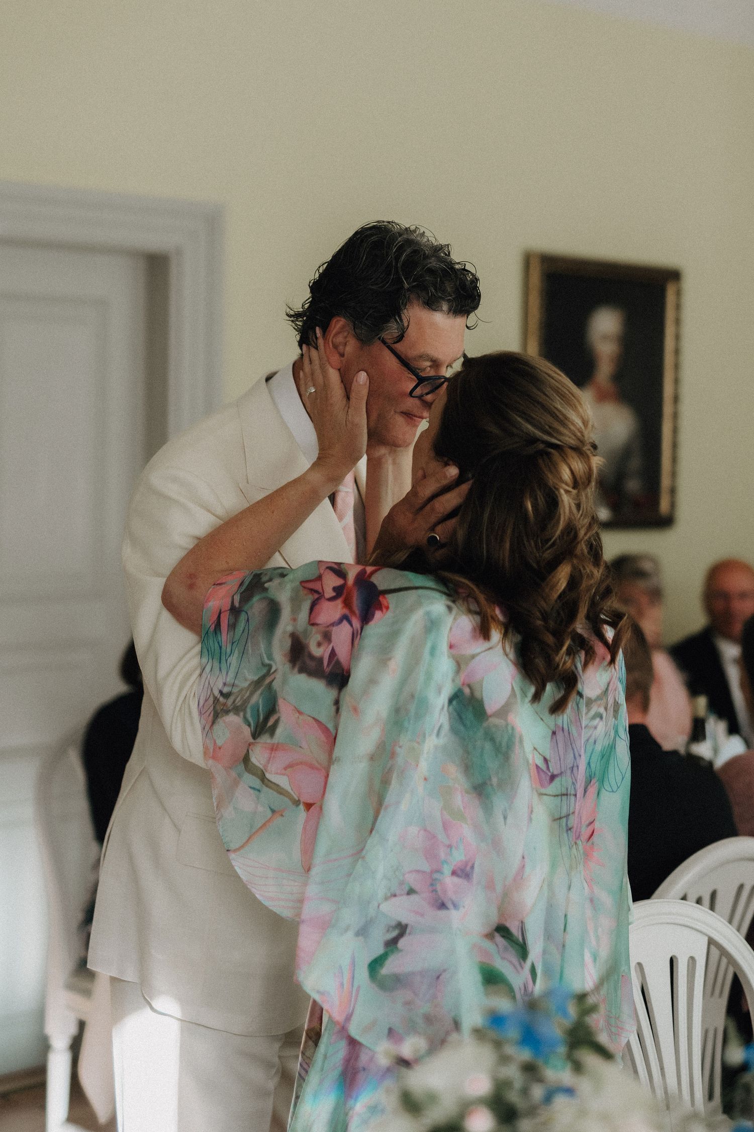 Man in white suit shares a tender kiss with woman in floral dress at wedding reception.