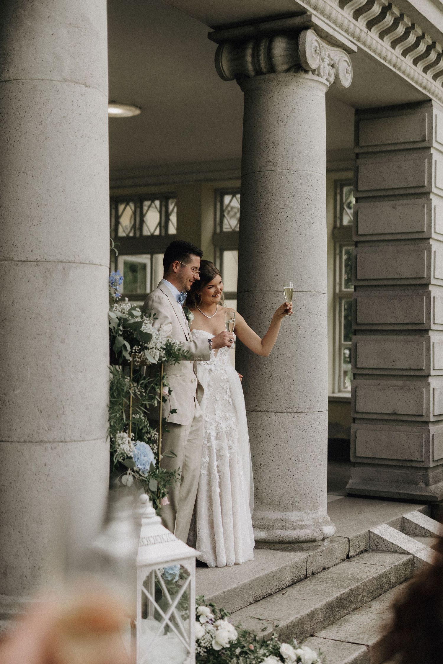 Bride and groom share a champagne toast on the steps of a classical building with tall columns.