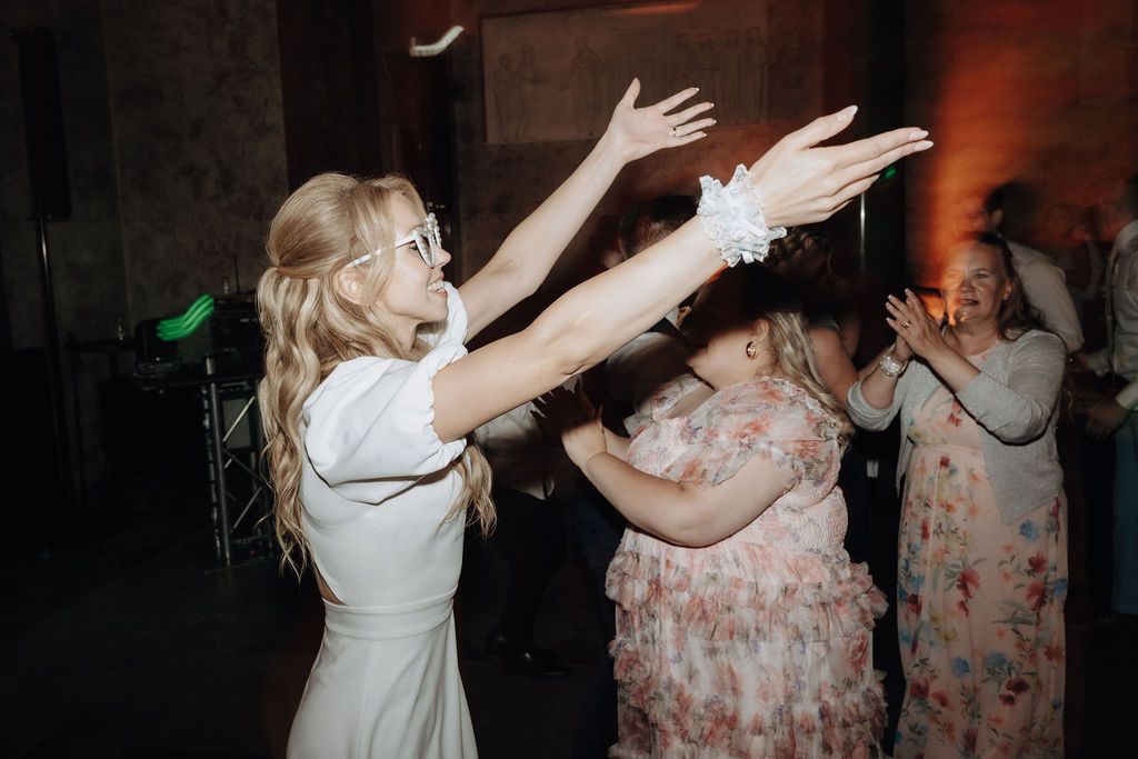 Blonde bride in white dress raises arms joyfully while dancing at wedding reception party.