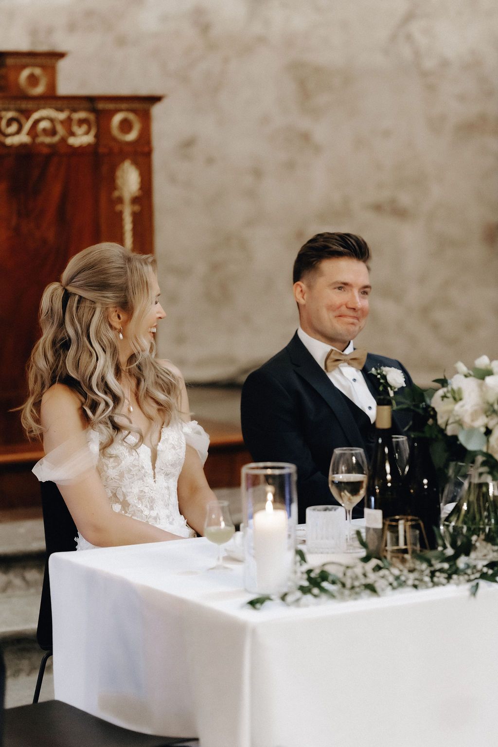Smiling bride and groom sit together at their elegantly decorated wedding head table.