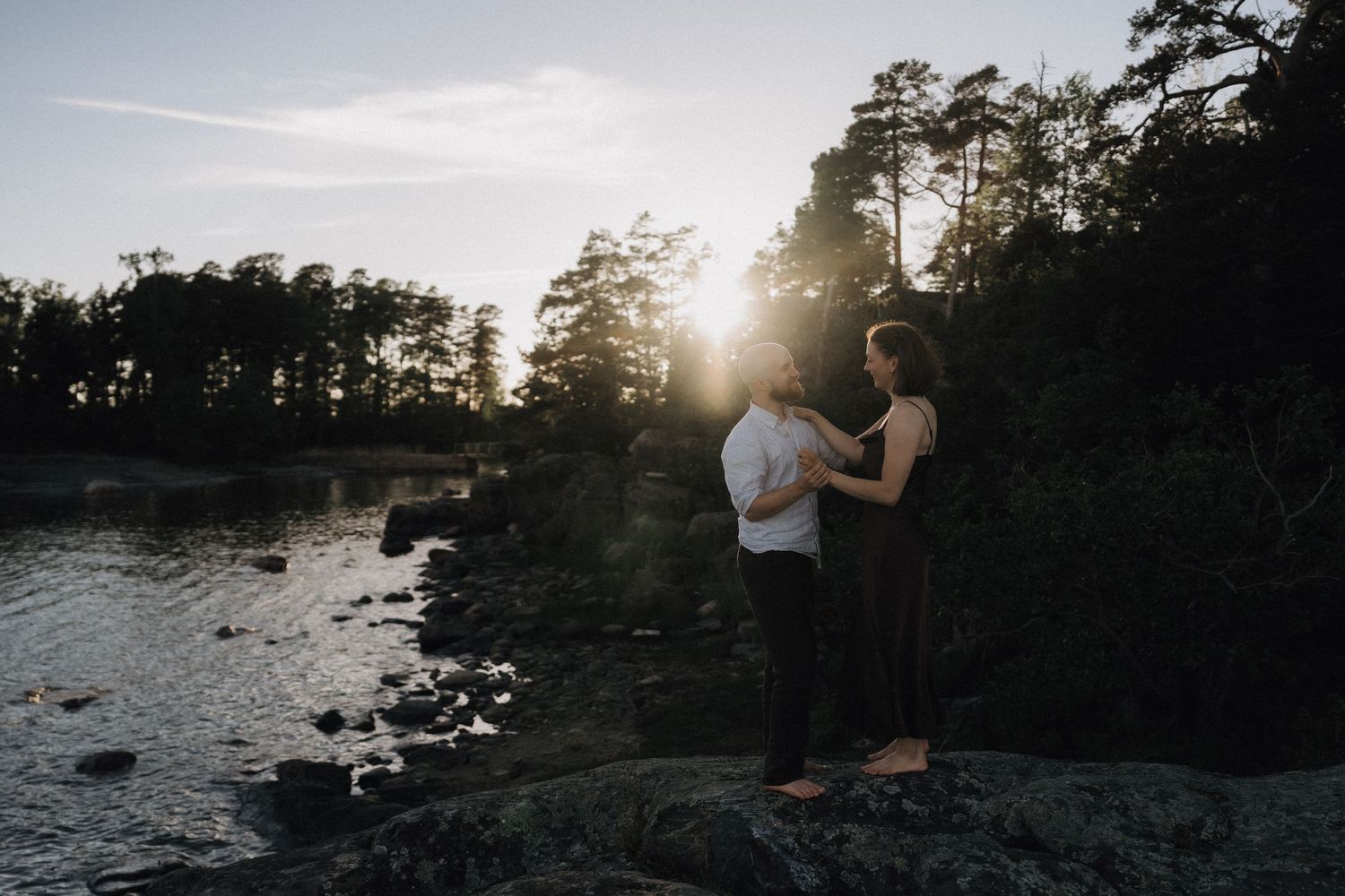Couple standing on rocks by a forest lake at sunset, facing each other in conversation.
