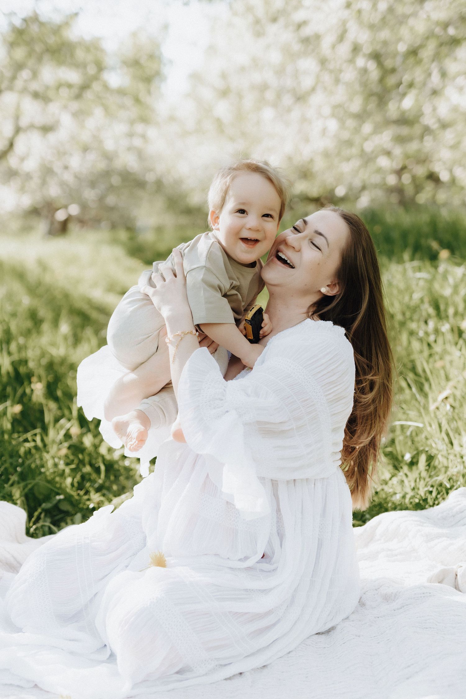A mother in a white dress sits on a blanket in a blooming garden, joyfully lifting her laughing baby.