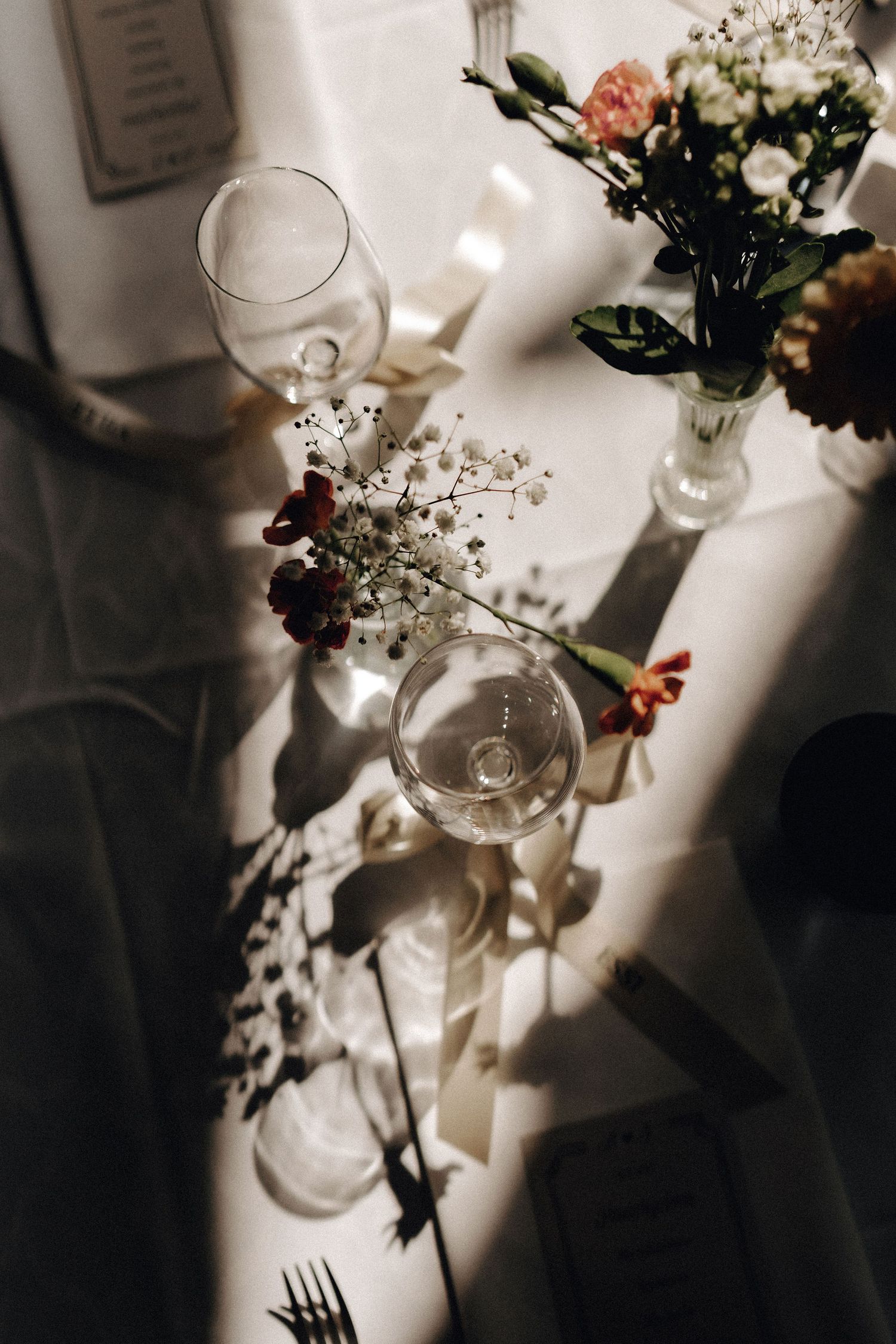 Elegant wedding reception table with glassware, floral arrangements, and soft sunlight casting shadows.