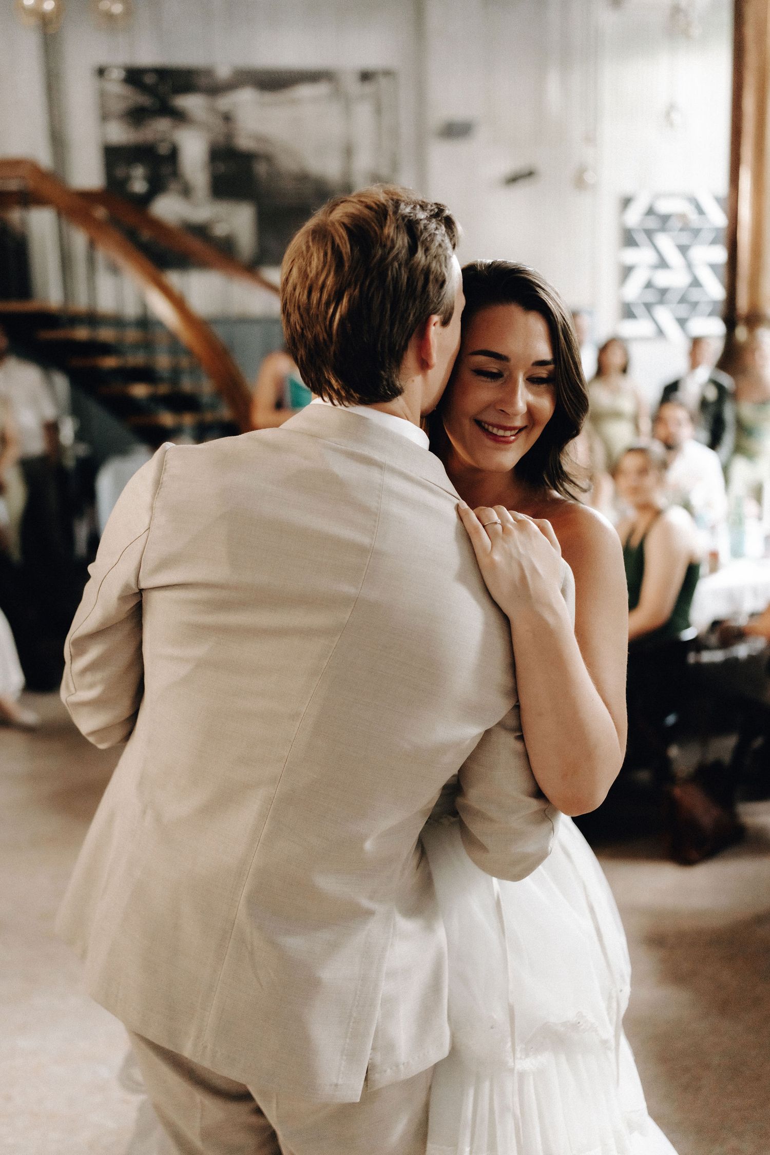 Smiling bride in white strapless gown embraces groom during first dance at wedding reception.