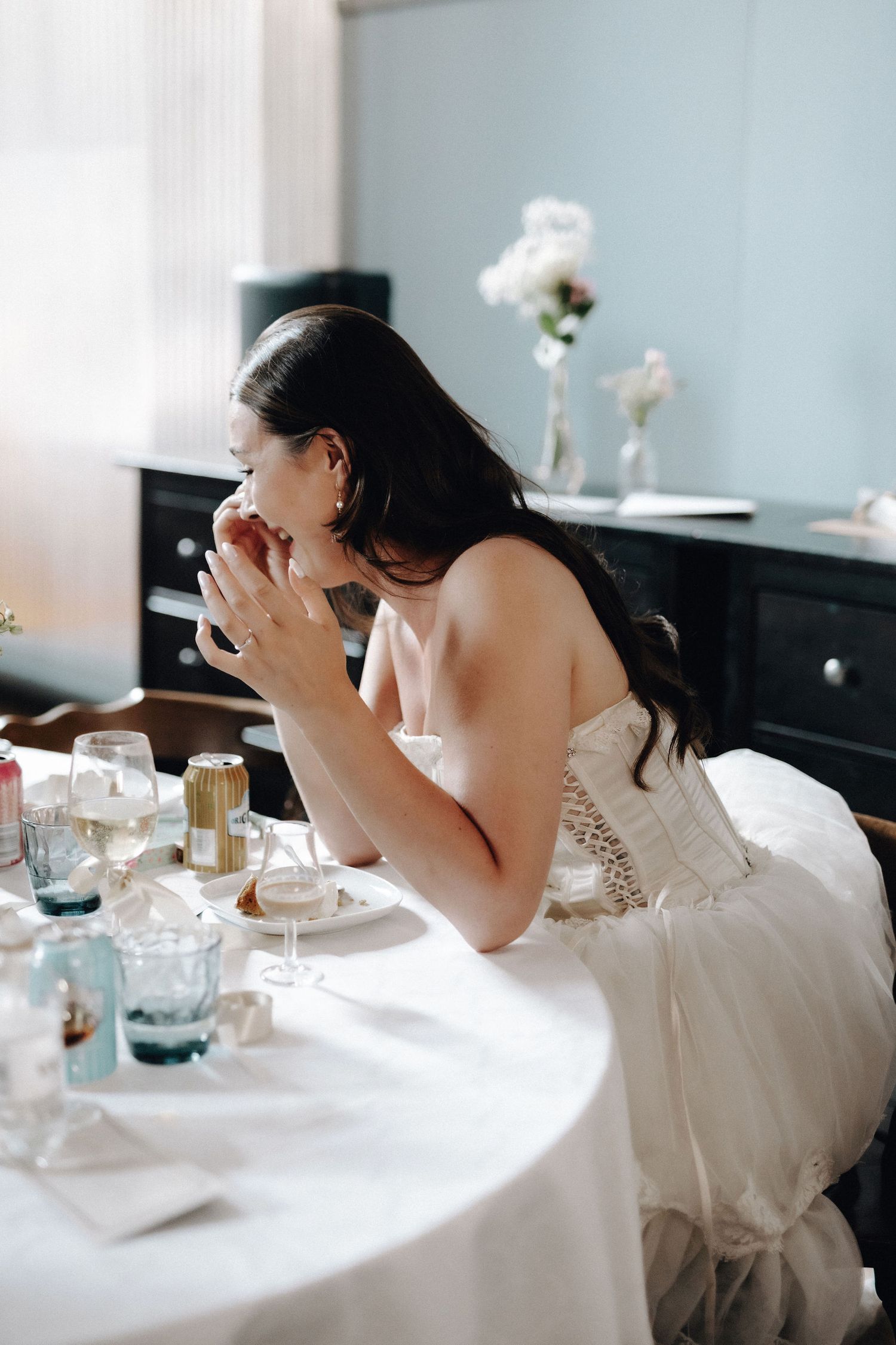 Emotional bride in white corset gown covering her face at wedding reception table.