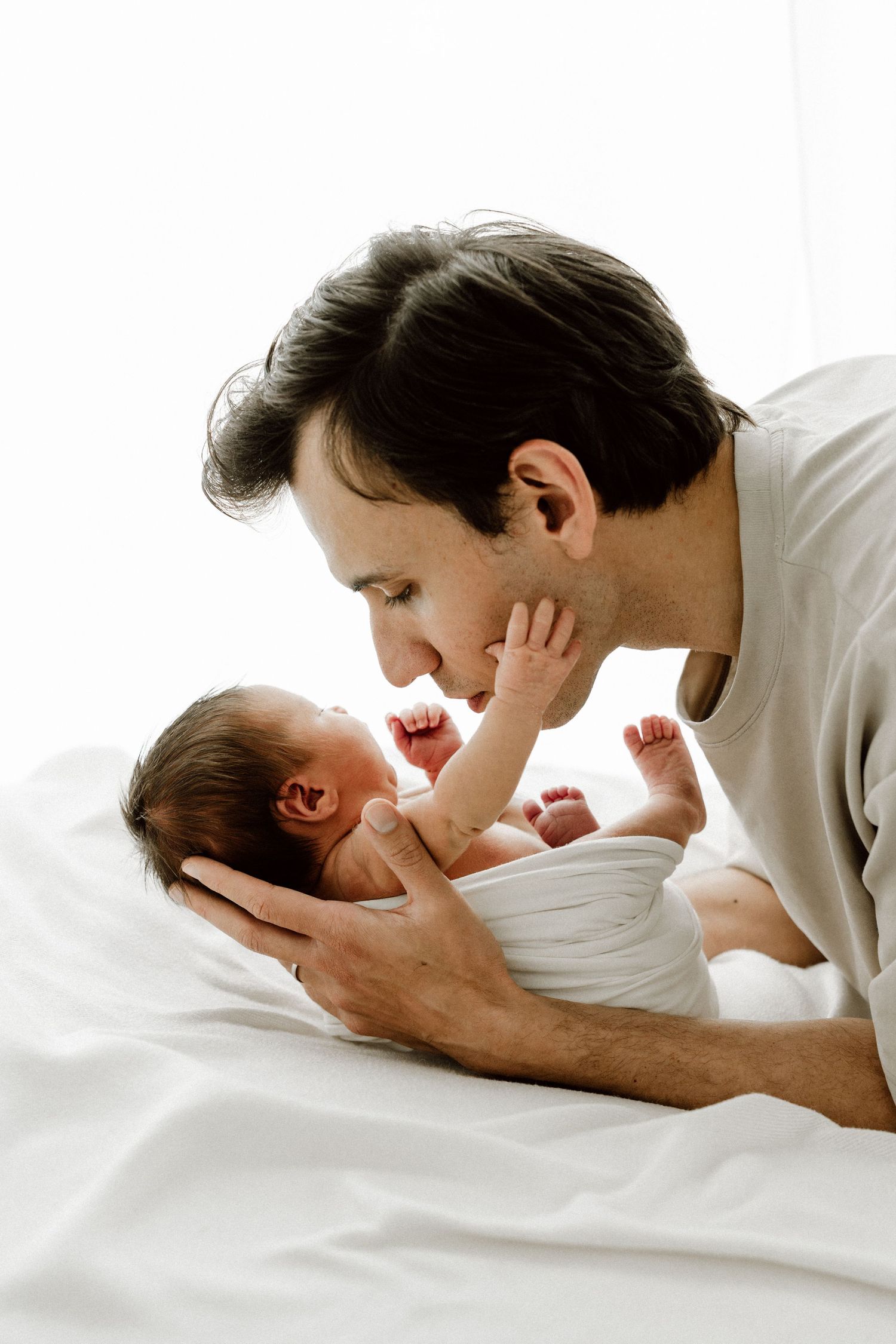 Dark-haired father leaning over white bed gazing lovingly at his newborn baby.