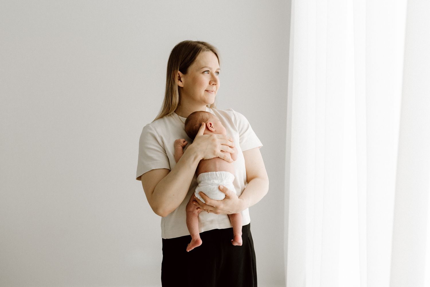 Young mother in white shirt holding sleeping newborn baby near bright window.