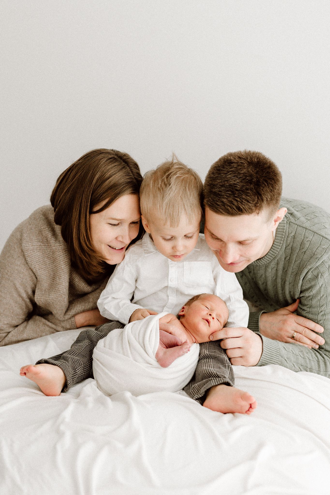 A happy family of four with parents and toddler son gathered around their swaddled newborn baby.