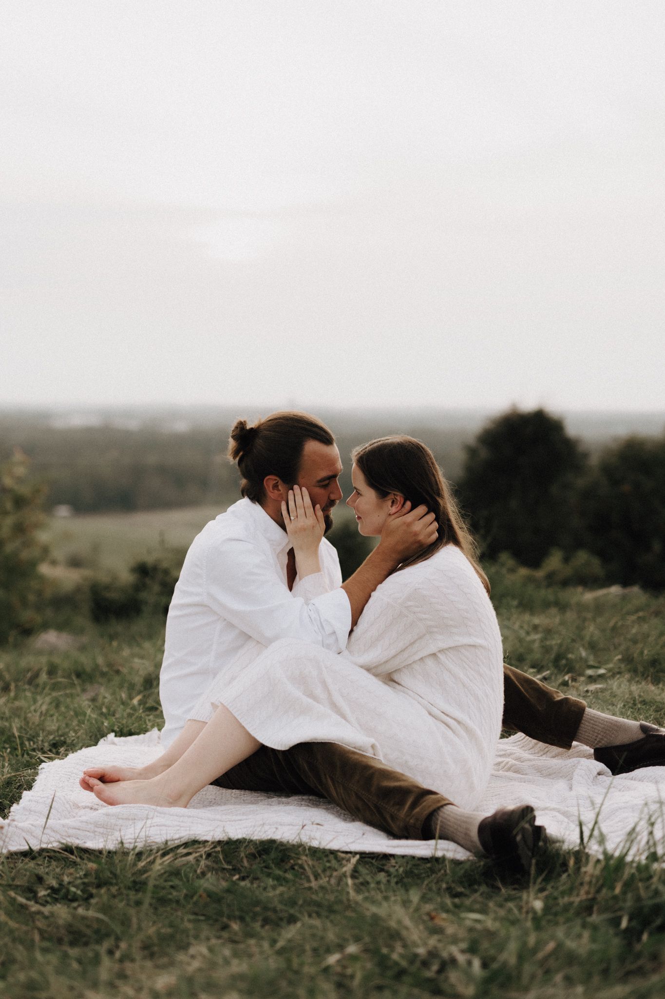 Couple sitting closely on a white blanket outdoors, gently holding each other's faces.