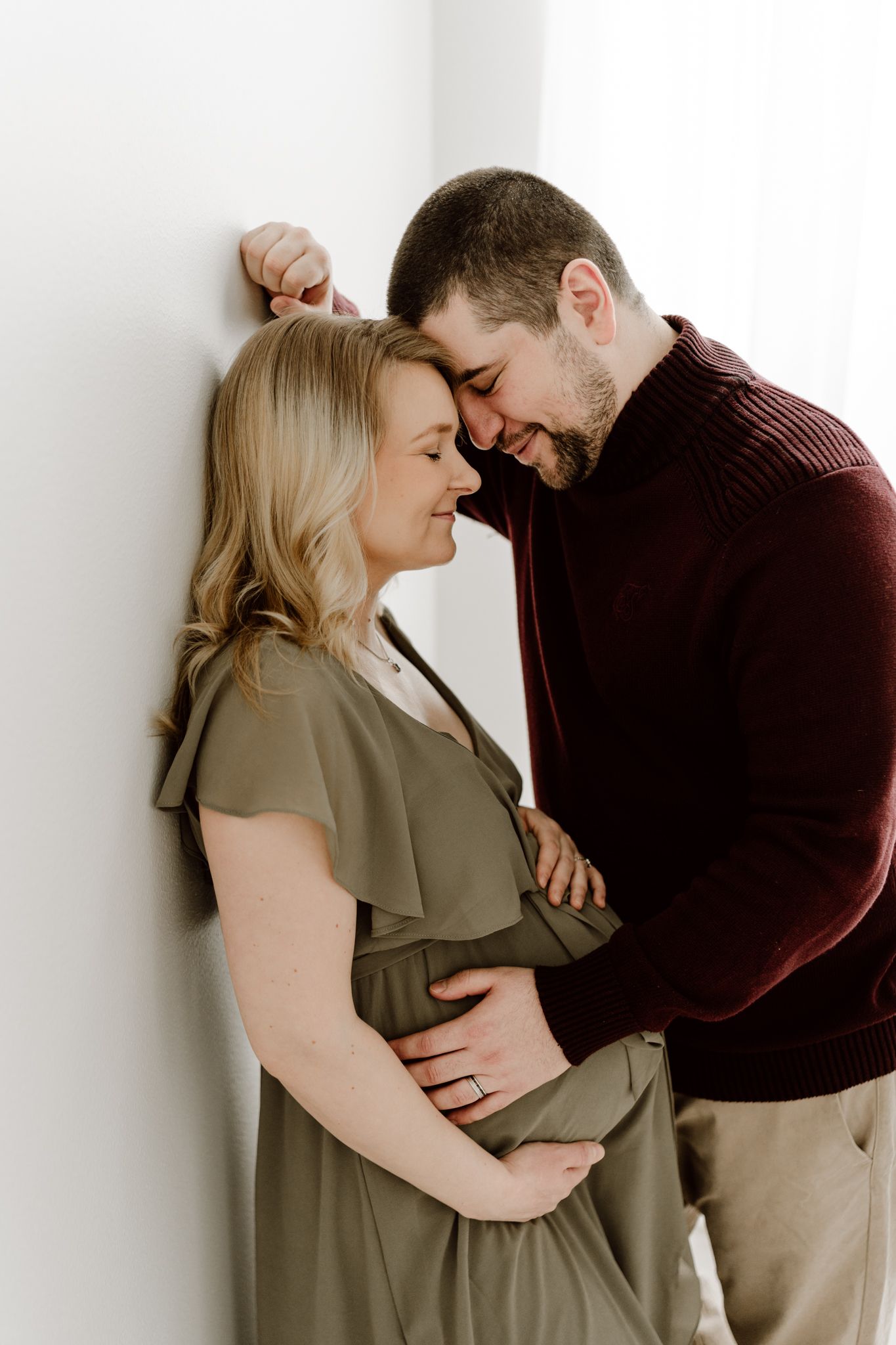 Expecting couple sharing intimate moment against white wall, woman in olive dress, man in burgundy sweater, foreheads touching.