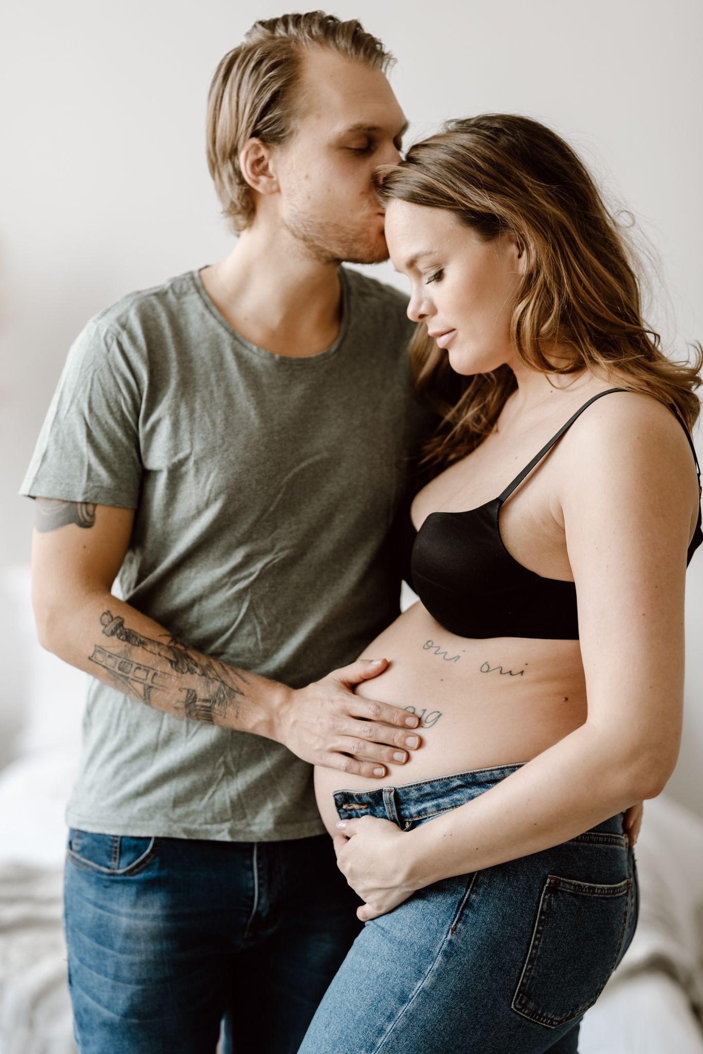 Man kissing pregnant woman on temple while holding her tattooed baby bump indoors.