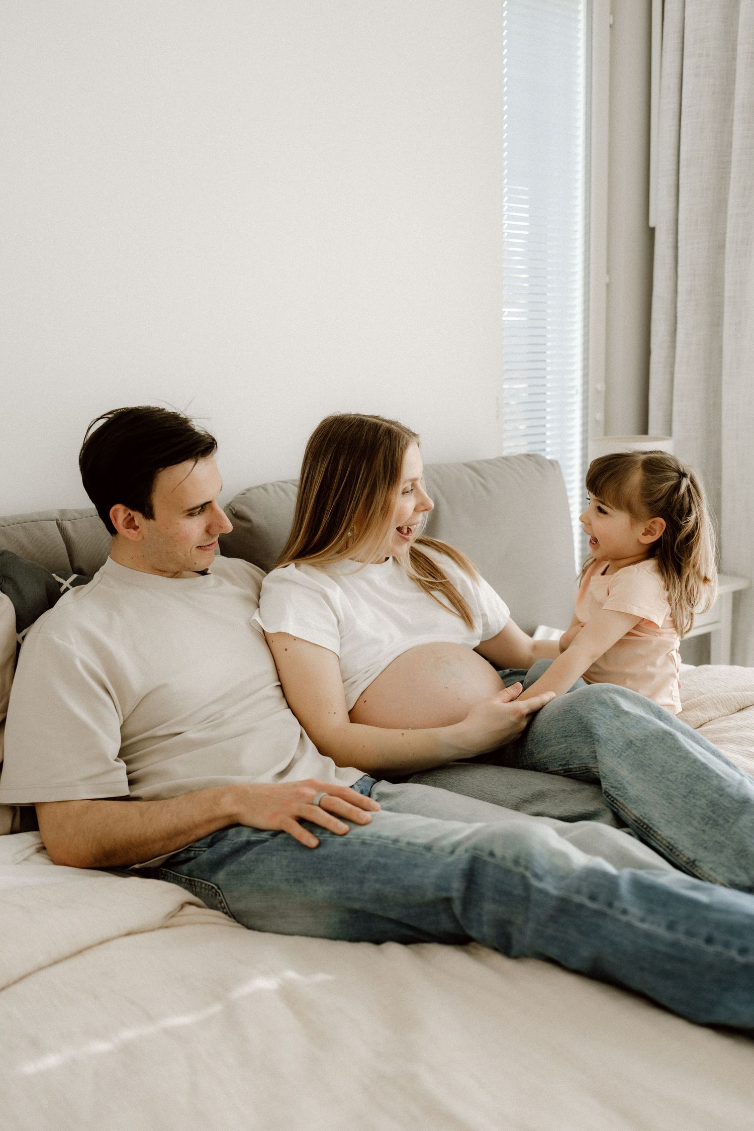 Family relaxing on sofa with father, pregnant mother, and young daughter chatting together.