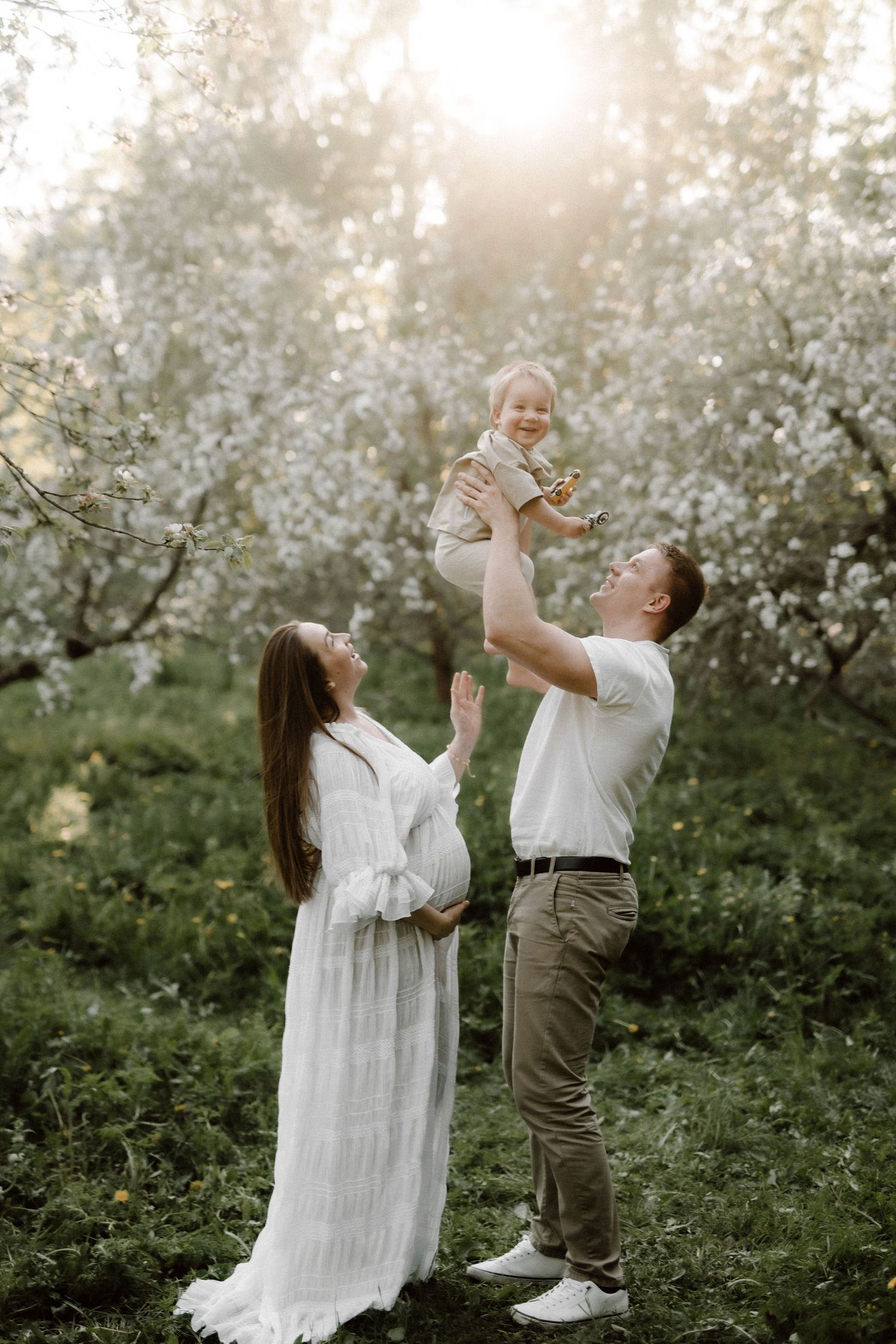 Joyful family in blooming orchard with father lifting laughing toddler as pregnant mother in white dress watches.