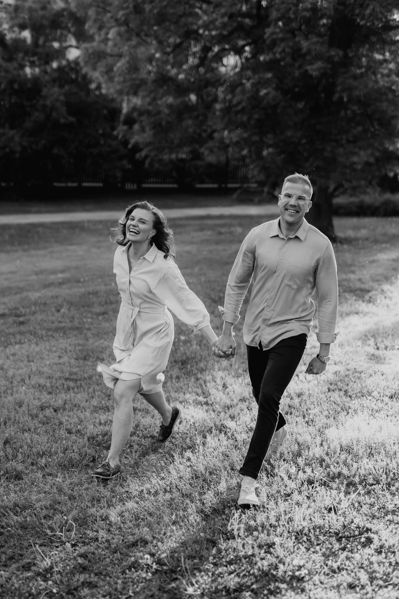 Black and white photo of laughing couple holding hands running through a park together.